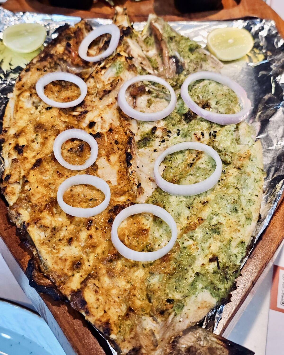 A whole fish grilled under a green marinade, laid out on a tray with rings of raw onion.