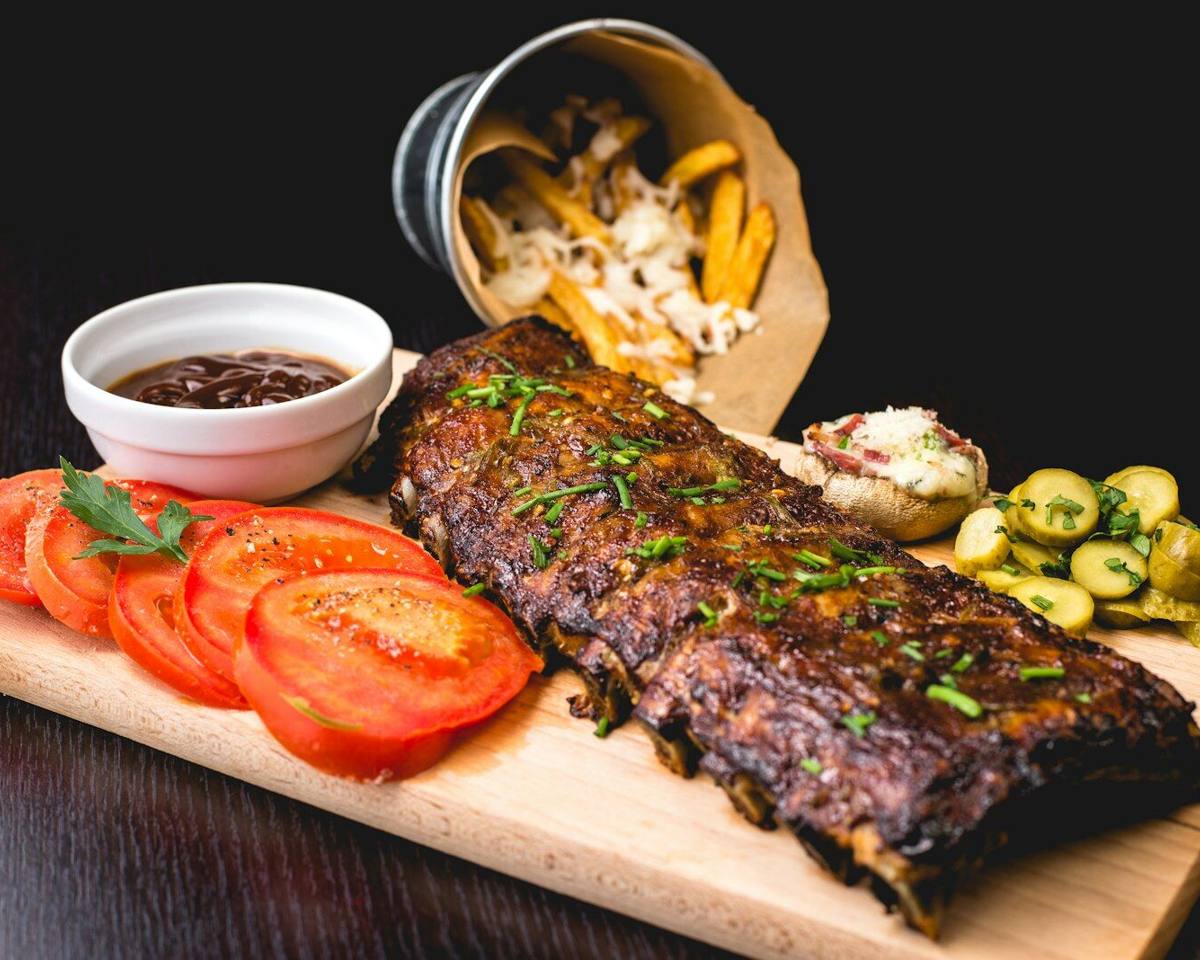 A rack of smoked ribs on a serving board with pickles and sliced tomato.