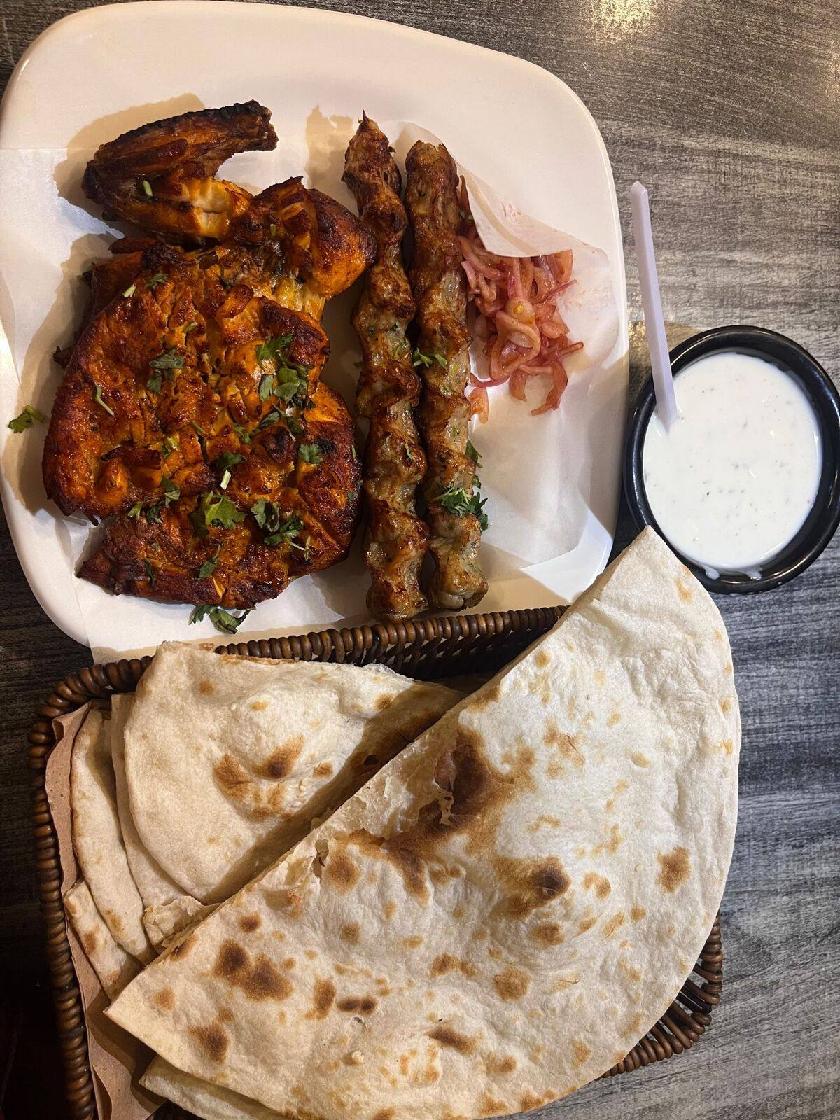 Grilled kebabs and skewers on a plate beside a basket of warm flatbread and a small pot of raita.