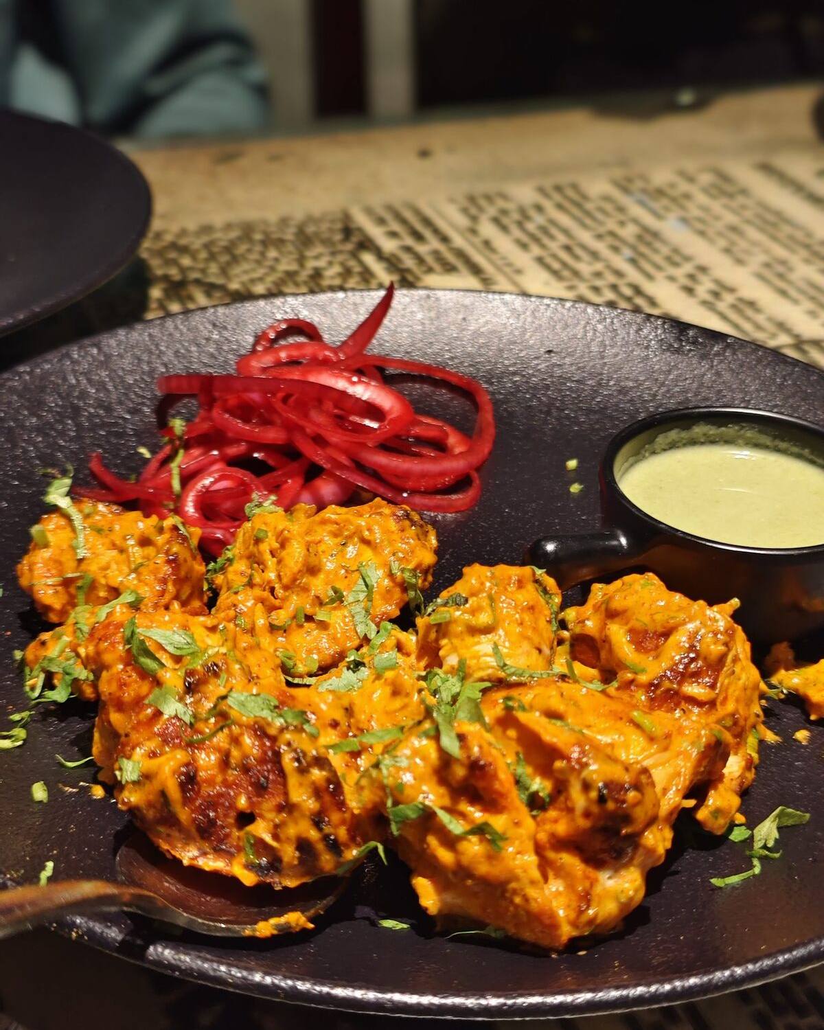 Charred tikka pieces on a dark platter with curls of red onion and a bowl of green chutney.
